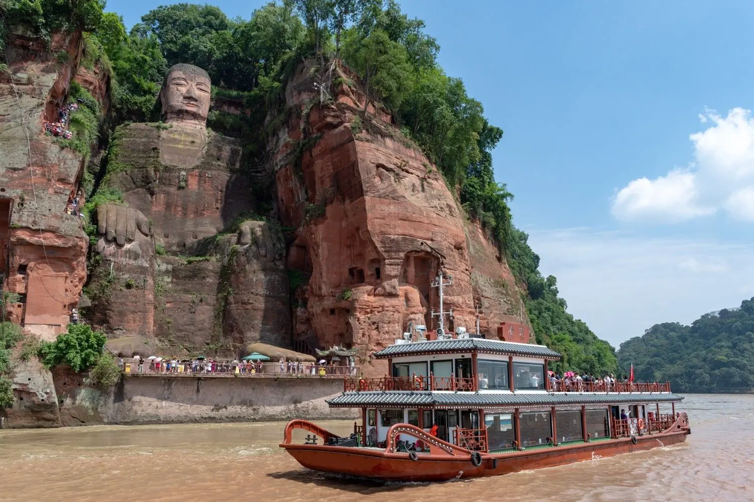 Leshan Giant Buddha in Sichuan, China, viewed from a sightseeing boat on the Min River, showcasing the world's largest stone Buddha carved into a red sandstone cliff and a UNESCO World Heritage Site.