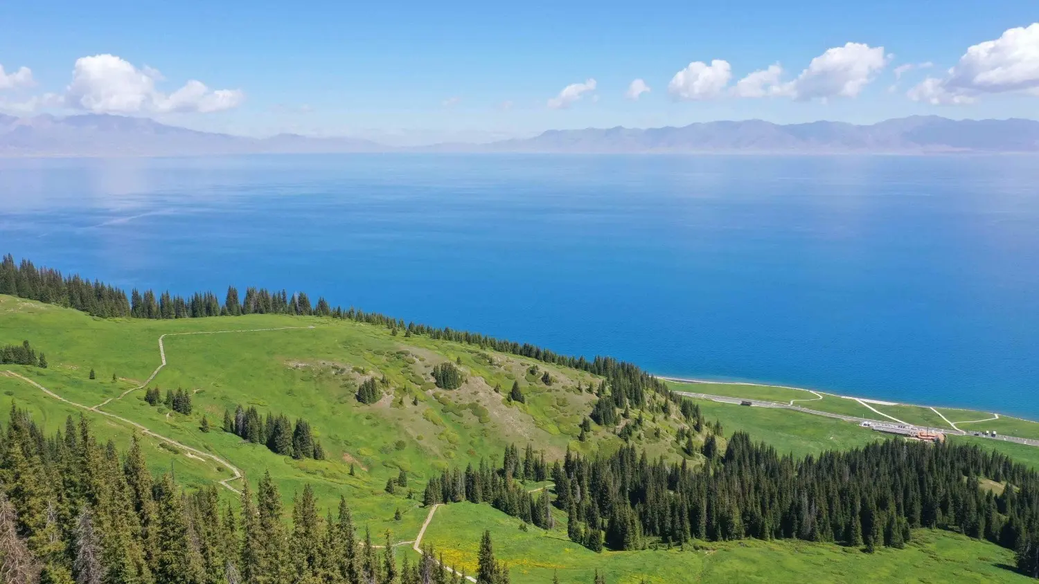 Panoramic view of Sayram Lake in Xinjiang, China, featuring crystal-clear blue waters, rolling green grasslands, and breathtaking alpine scenery along the famous Silk Road.