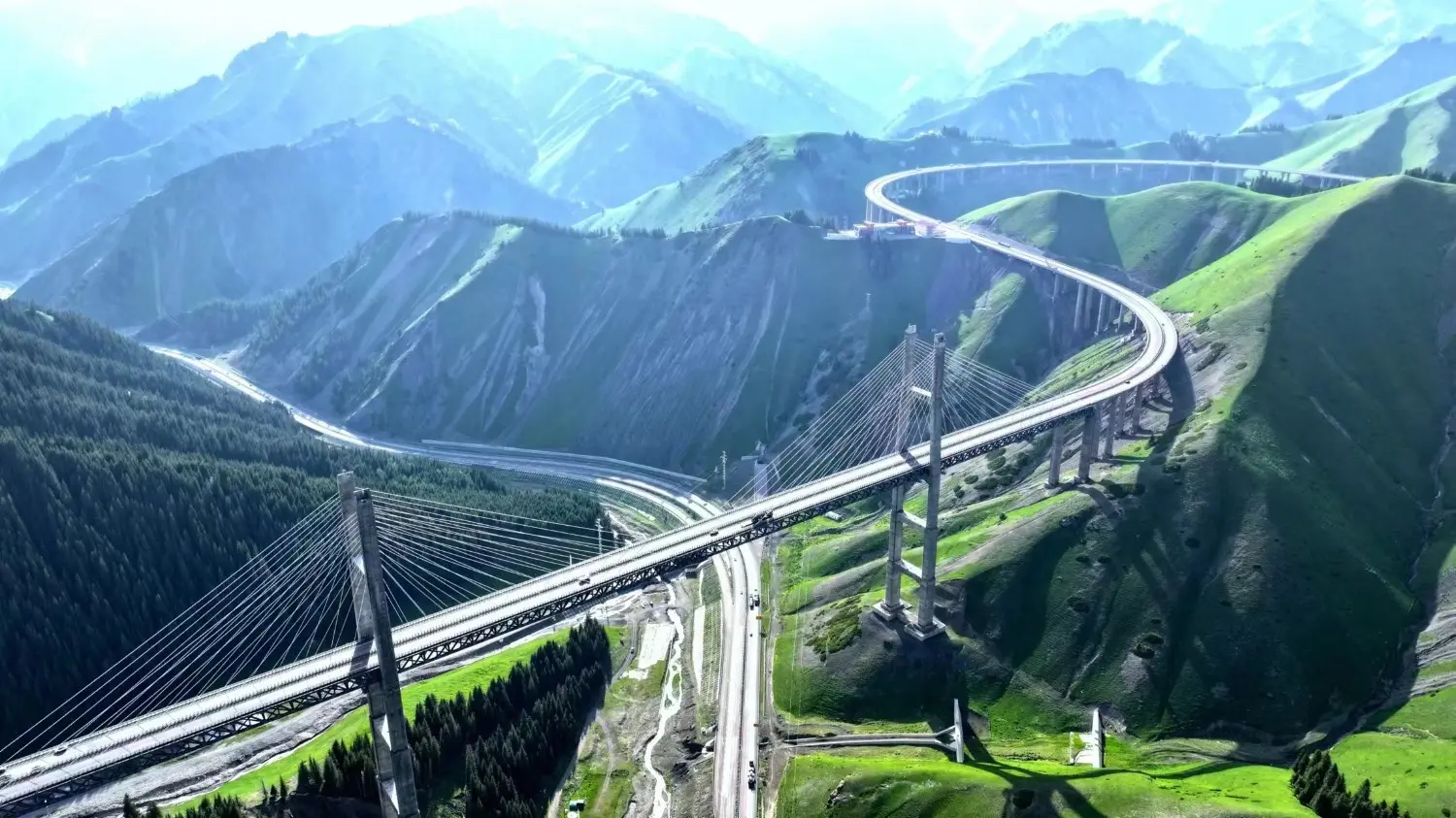 Aerial view of Guozigou Bridge in Xinjiang, China, winding through lush green mountains and deep valleys, showcasing one of the country's most impressive engineering landmarks along the scenic Ili Highway.