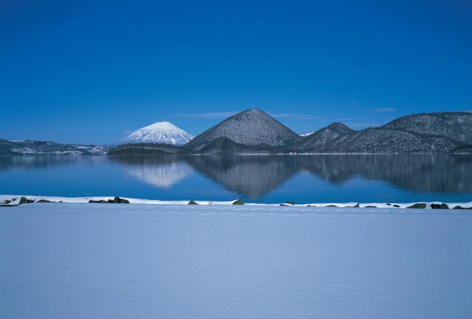 Lake Toya in Hokkaido, Japan, featuring a serene snow-covered lakeshore, crystal-clear waters reflecting volcanic mountains, and a peaceful winter landscape. One of Hokkaido's most scenic destinations, renowned for its natural beauty, tranquil atmosphere, and year-round outdoor experiences.
